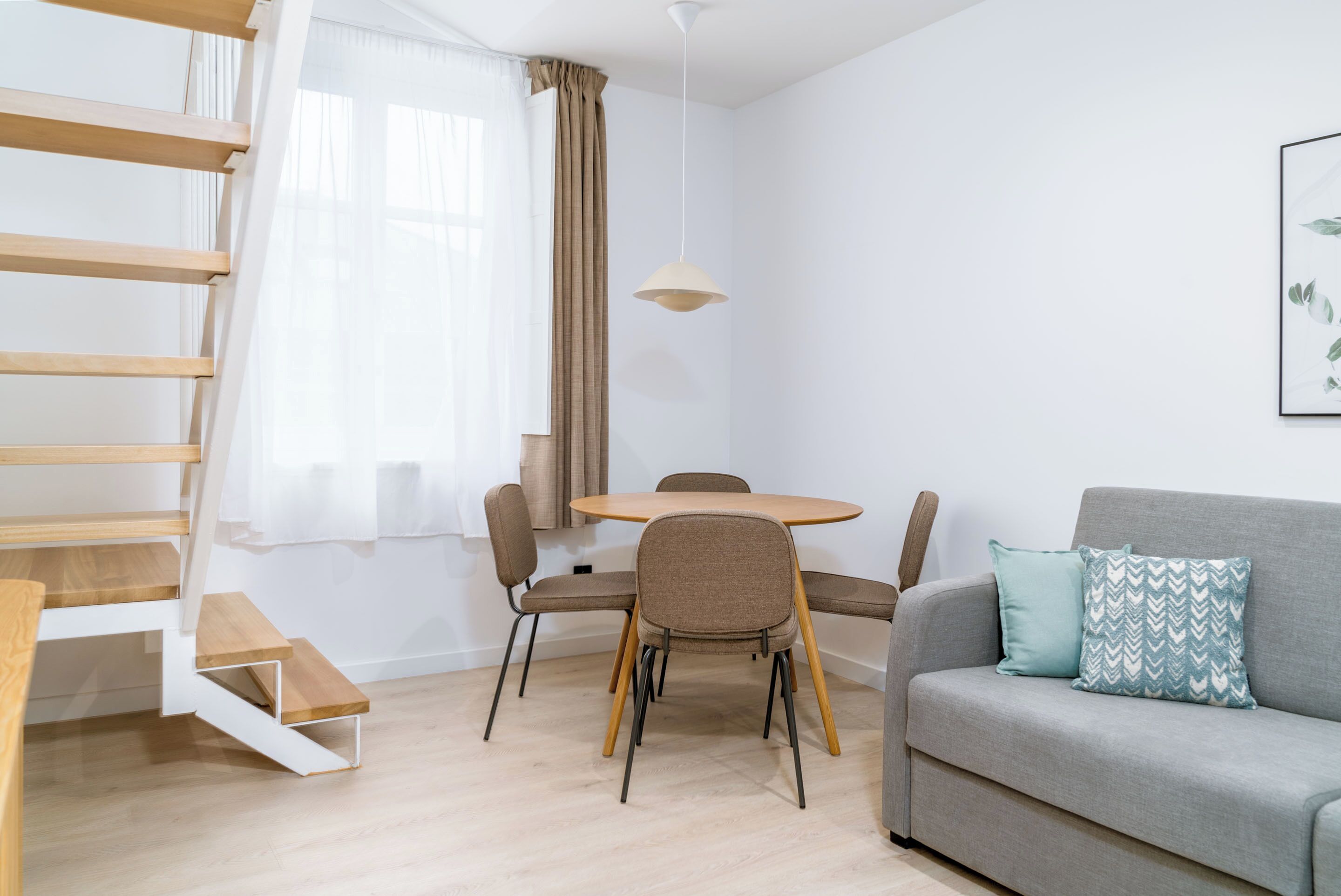 A modern living room with a staircase, a round dining table, and a grey sofa with patterned cushions.