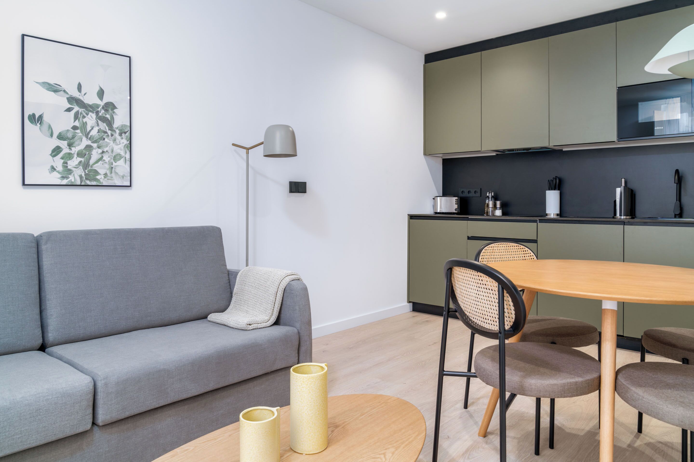 Una sala de estar moderna con un sofá gris, una mesa de comedor de madera y una cocina con armarios verdes y un microondas.