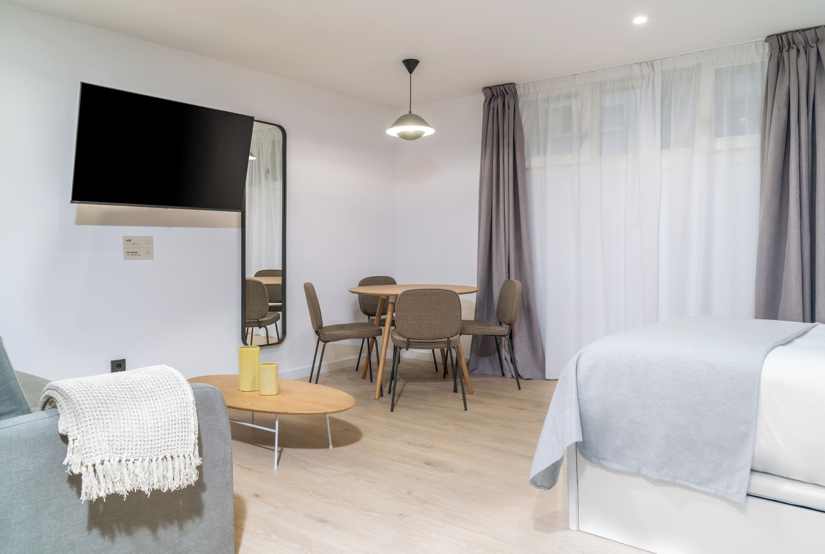 A modern hotel room with a sofa, a bed, a dining table with chairs, a flat-screen TV mounted on the wall, and a ceiling light.