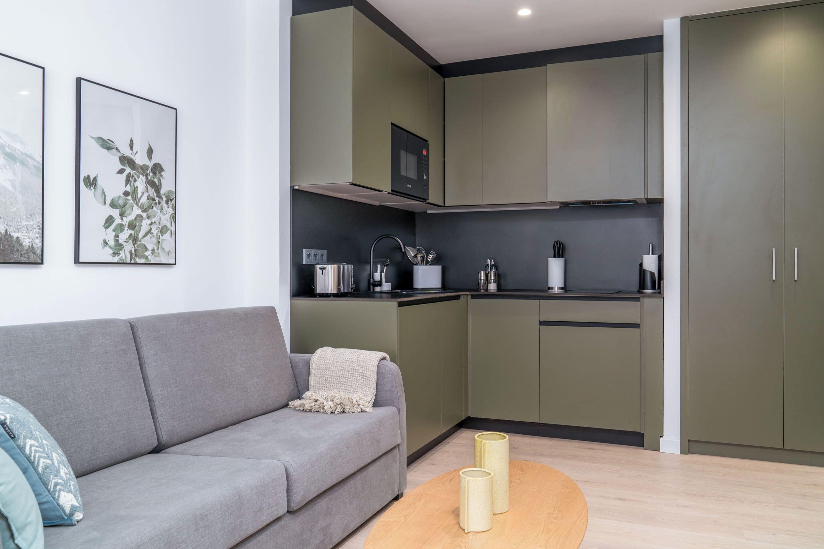 A modern kitchen with green cabinets, a grey sofa, and a wooden coffee table.