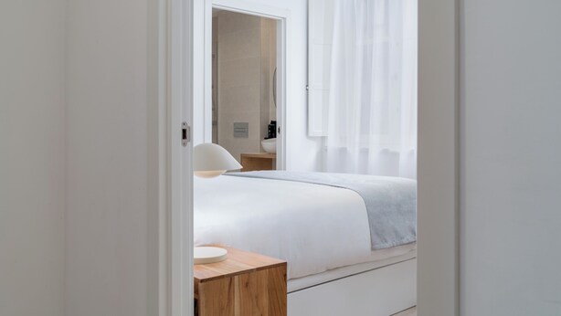 Quarto de hotel moderno com uma cama bem arrumada, uma mesa de cabeceira e um banheiro visível através de uma porta aberta.