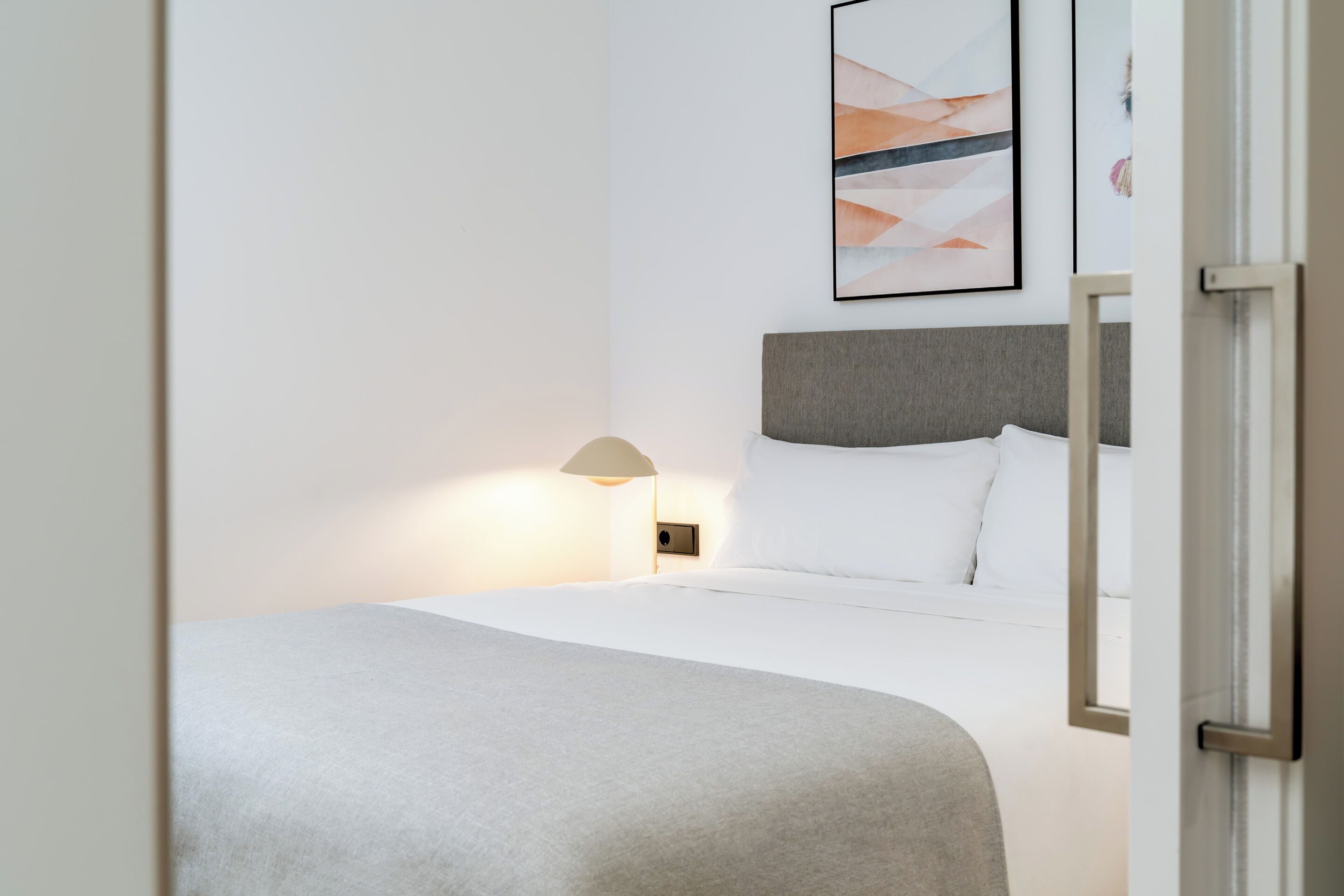 A neatly made bed with a grey headboard, a bedside lamp, and a framed abstract artwork on the wall.