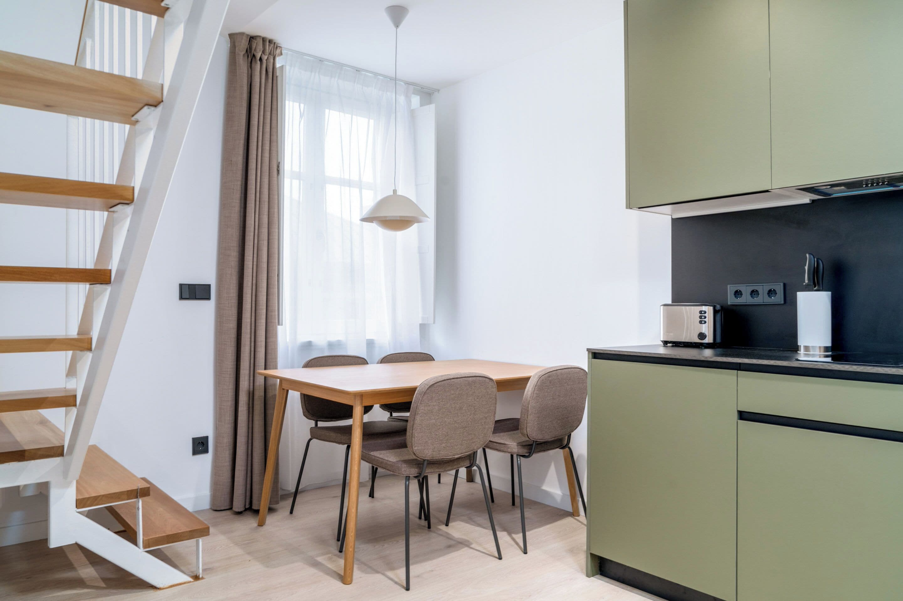 A modern kitchen with a dining area, a staircase, and a window with curtains.