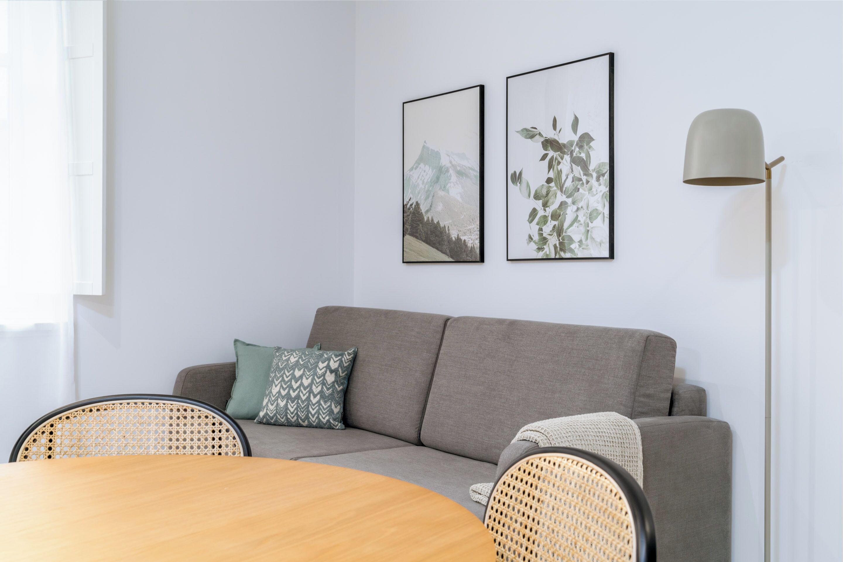A modern living room with a grey sofa, two framed pictures, a floor lamp, and a round wooden table with rattan chairs.
