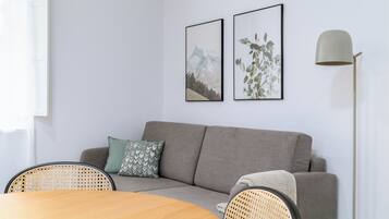 A modern living room with a grey sofa, two framed pictures, a floor lamp, and a round wooden table with rattan chairs.