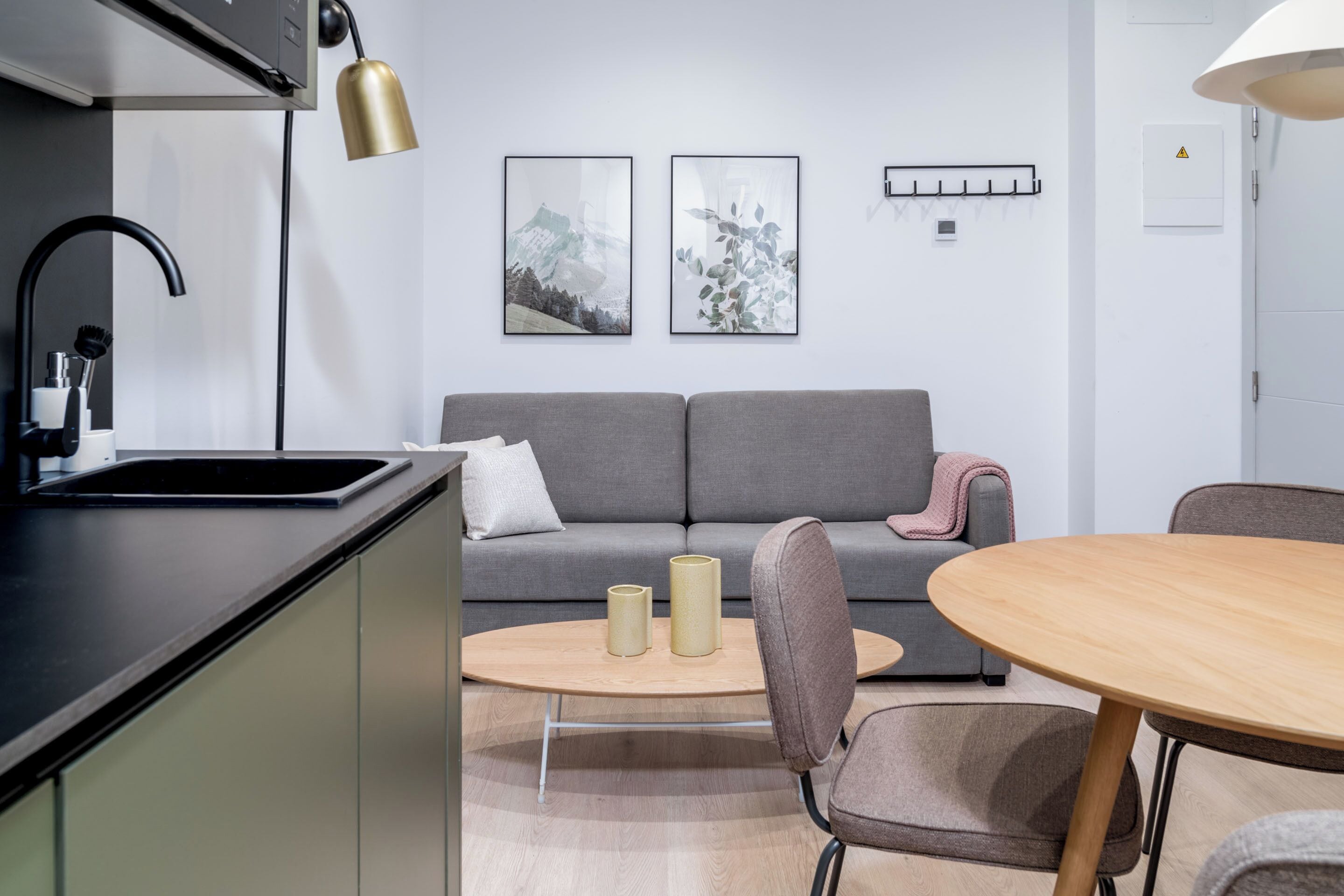 Una cocina moderna con encimera negra, un lavabo y un grifo negro. Hay un sofá gris con una manta rosa y dos cuadros en la pared. En primer plano, una mesa redonda de madera con dos sillas.