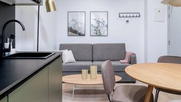 A modern kitchen with a black countertop, a sink, and a black faucet. There is a grey sofa with a pink throw blanket and two framed pictures on the wall. A round wooden table with two chairs is in the foreground.