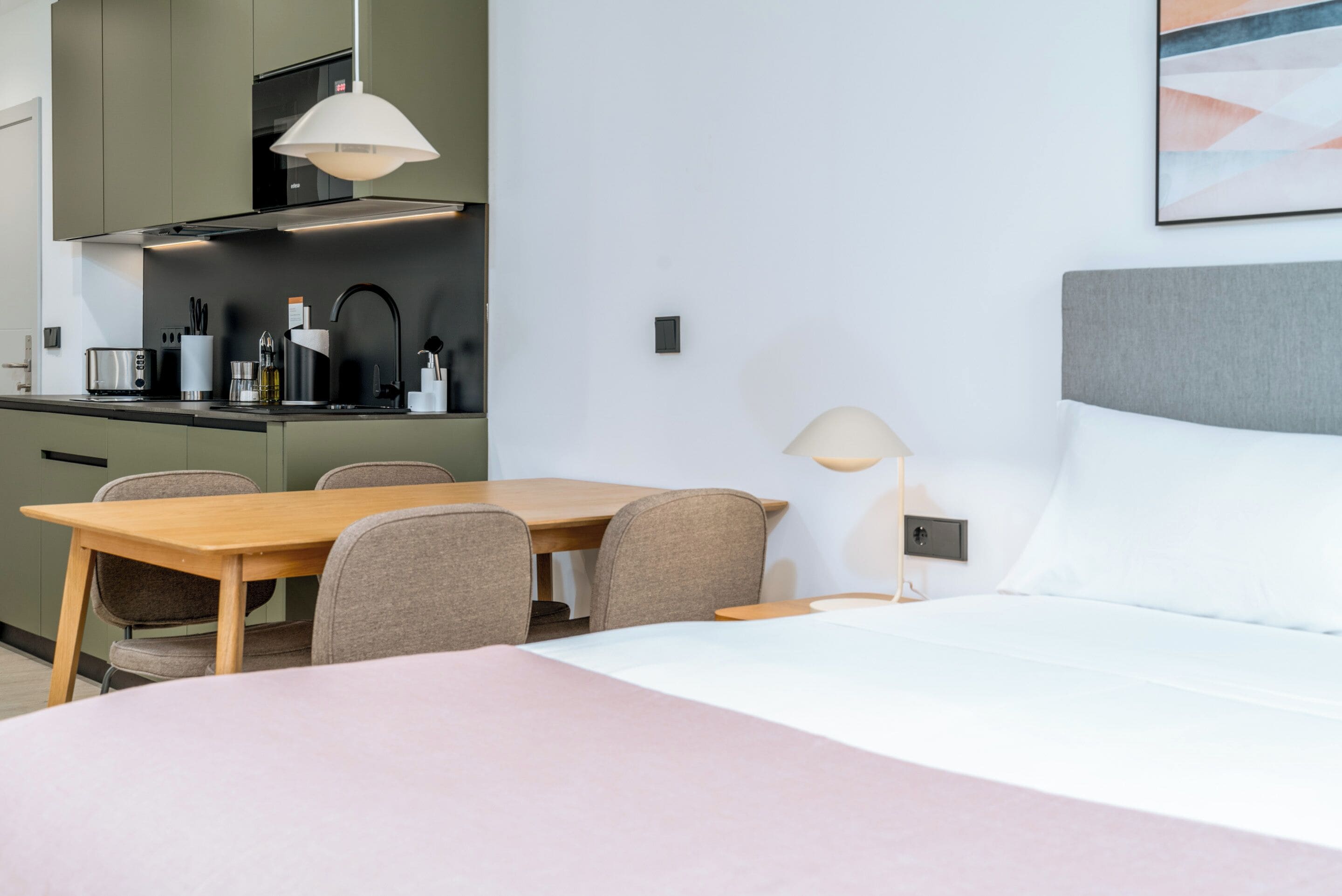 Una habitación de hotel moderna con cama, mesita de noche, lámpara y una zona de kitchenette con mesa y sillas.