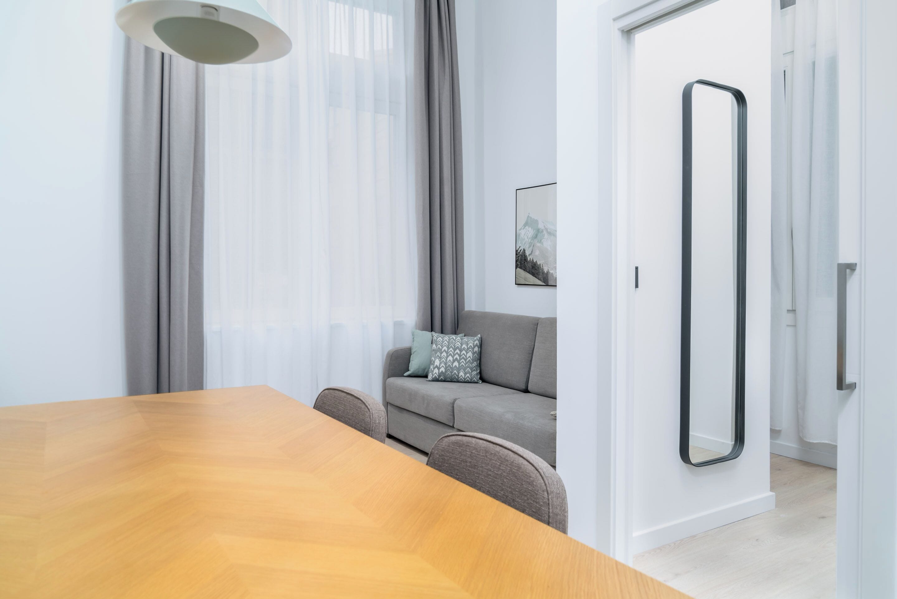 A modern living room with a wooden dining table, grey sofa, and a large mirror.