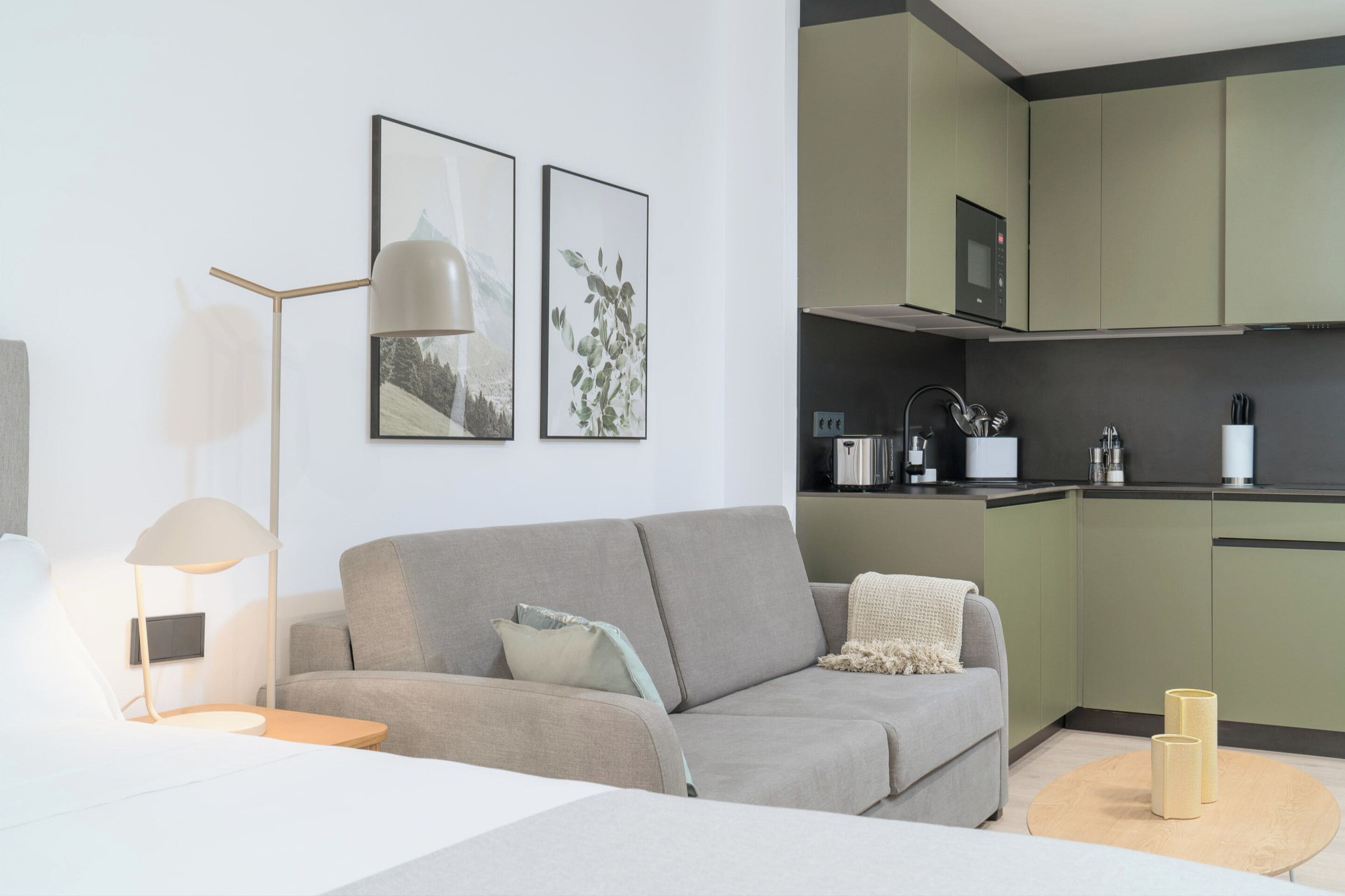 A modern living room with a grey sofa, a side table with a lamp, and a coffee table. There are two framed pictures on the wall, a kitchen area with green cabinets, and a microwave.