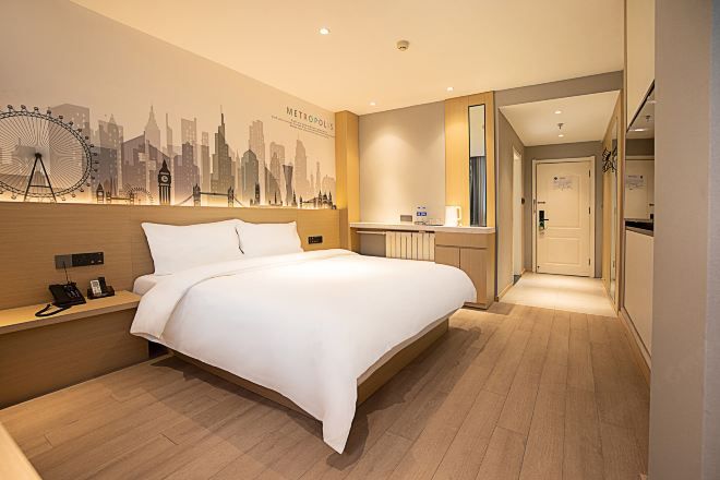 A hotel room with a bed, a nightstand, a mirror, a wall with a cityscape mural, and a hallway leading to a bathroom.