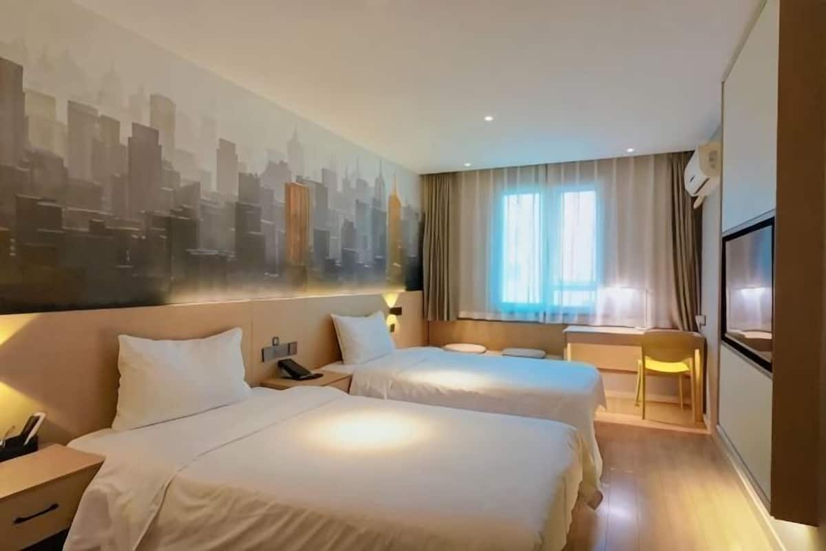 Habitación de hotel con dos camas, un mural de paisaje urbano, un ventanal con cortinas, un televisor y un escritorio con una silla.