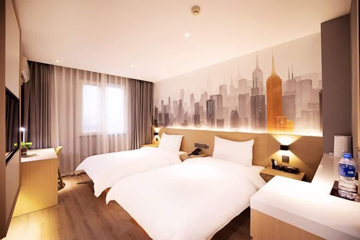 Habitación de hotel con dos camas, un mural de la ciudad, un escritorio con teléfono y una mesita de noche con lámpara.
