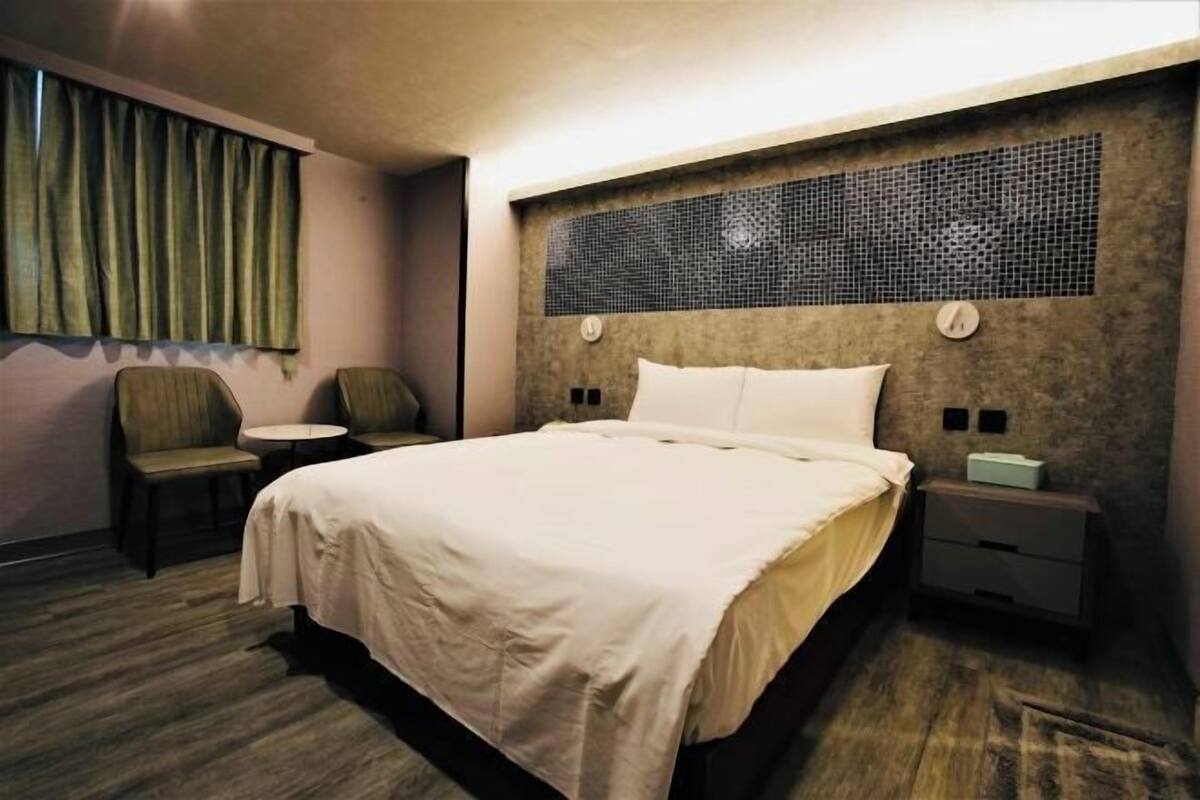 A hotel room with a bed, a small round table, two chairs, a nightstand with a lamp, and a decorative wall panel.