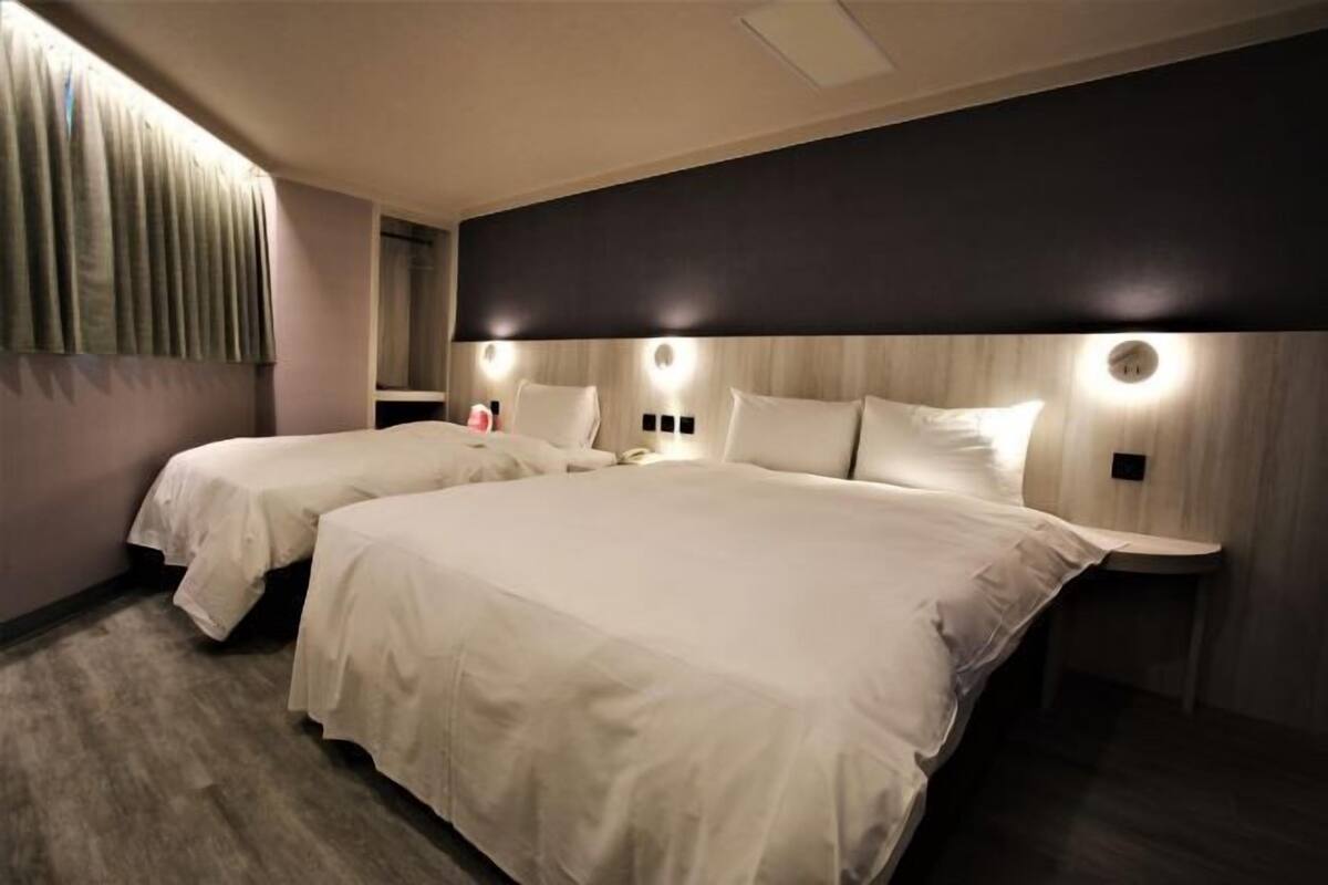 A hotel room with two beds, a large headboard with integrated lighting, a wooden accent wall, and a small bedside table.