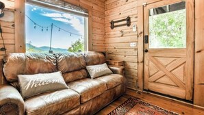 Interior de uma cabana de madeira com um sofá de couro, uma porta de madeira e uma janela com vista para o verde.