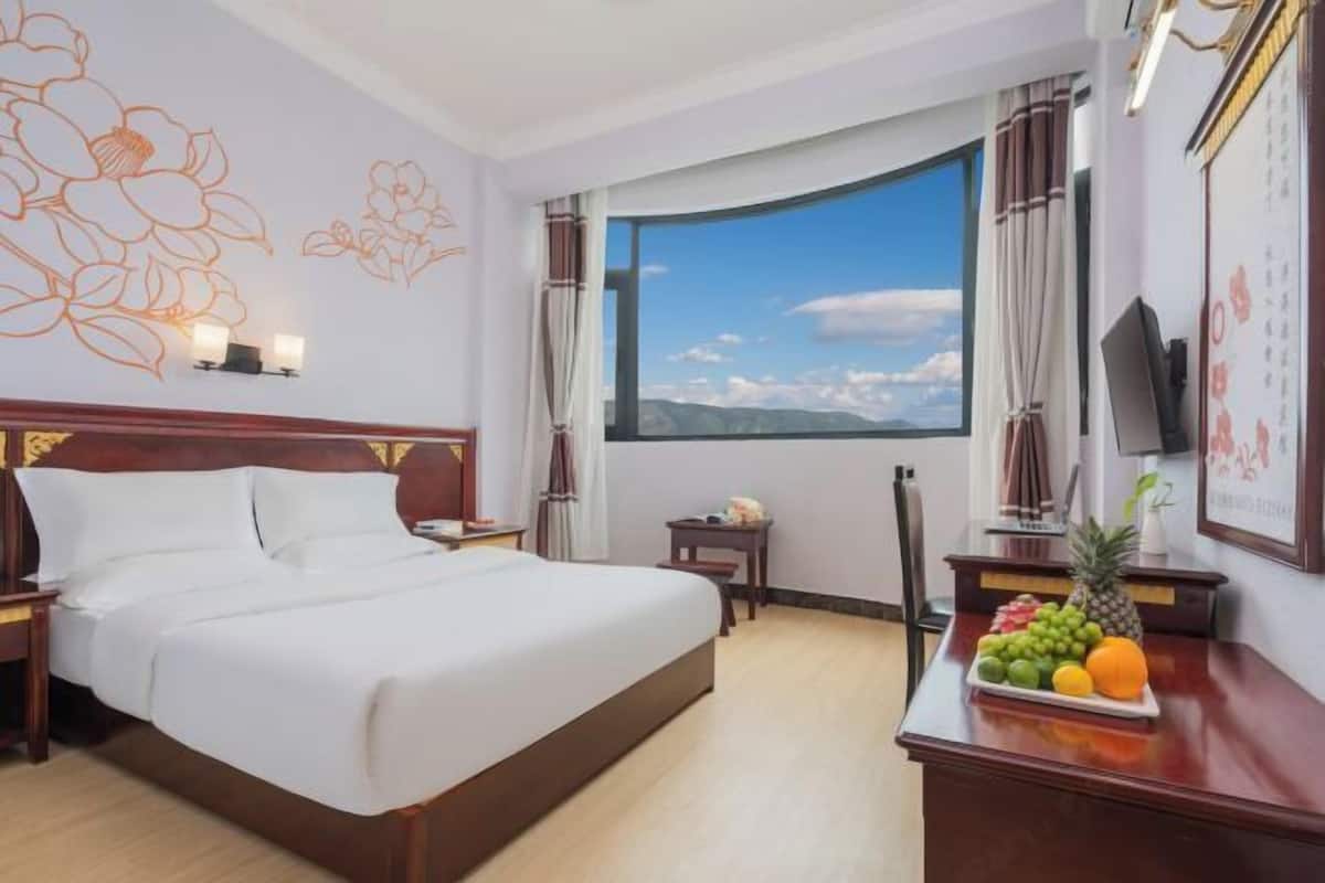 Ein Hotelzimmer mit einem großen Bett, einem Schreibtisch mit Obstschale, einem Fernseher und einem Fenster mit Blick auf die Berge.