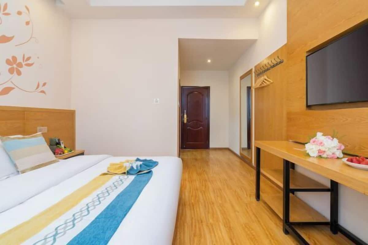Ein Hotelzimmer mit Bett, Holzboden, einem an der Wand montierten Fernseher und einem zur nächsten Zimmer führenden Gang.