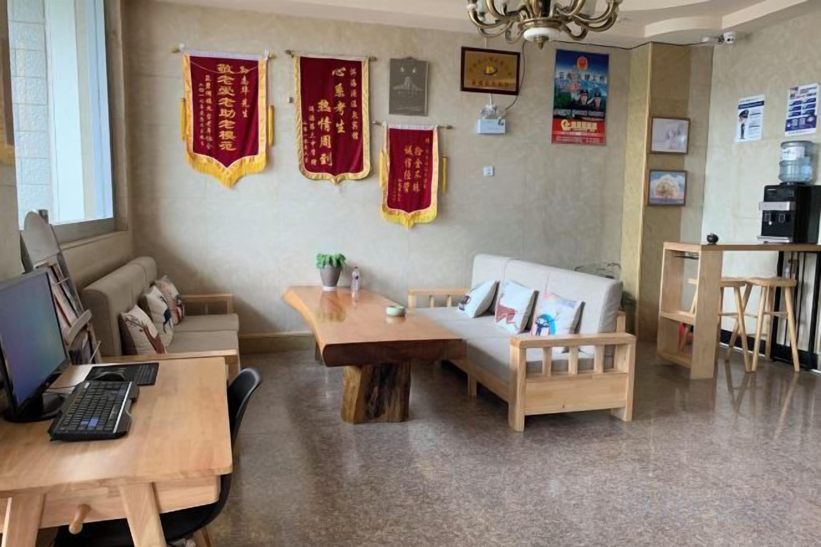 A room with a wooden desk, a white sofa, and a wooden dining table. There are red banners on the wall and a water dispenser on the right.
