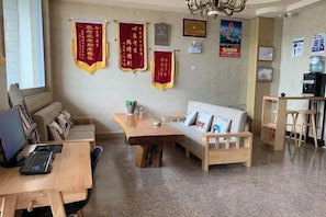 A room with a wooden desk, a white sofa, and a wooden dining table. There are red banners on the wall and a water dispenser on the right.