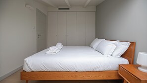 Un lit bien fait, avec du linge de lit blanc et des oreillers, une tête de lit en bois et une table de chevet surmontée d’une lampe blanche.