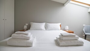 Un lit bien fait, avec du linge de lit blanc, et deux piles de serviettes pliées devant. On peut voir deux tables de chevet avec des lampes, une chaise grise et, au fond, une armoire.