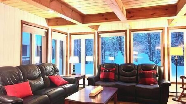 A cozy living room with a leather sofa and chairs, wooden beams on the ceiling, and large windows with a view of trees. - Family chalet and SPA near Mont Orford (Orford)