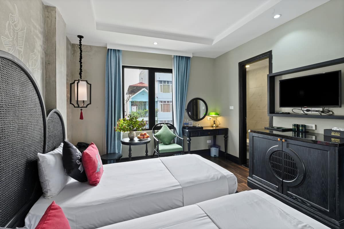Quarto de hotel com duas camas, televisão, escrivaninha com espelho, uma mesinha com planta e vista para edifícios.