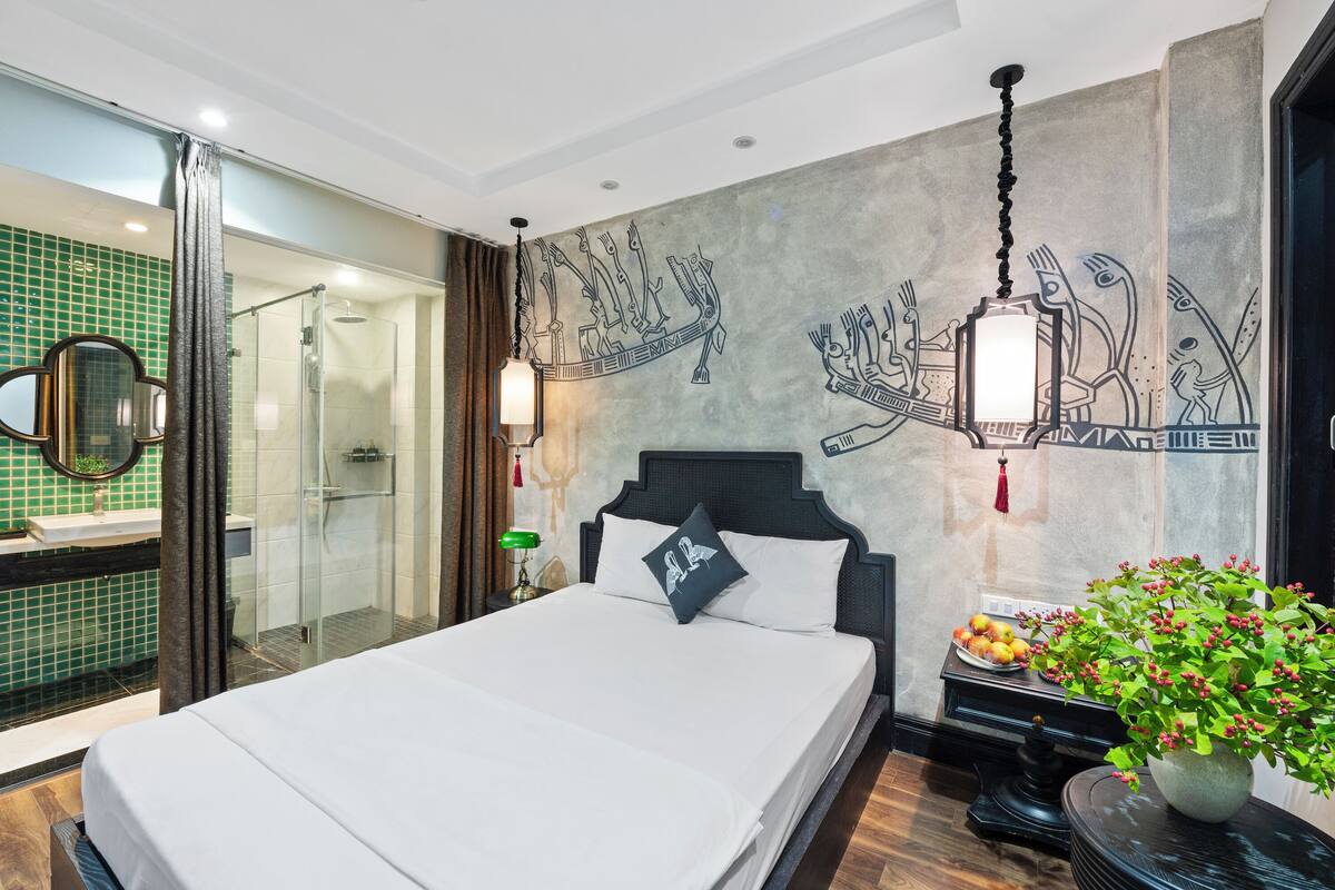 Quarto de hotel moderno com uma cama grande, box de vidro para o chuveiro, um mural decorativo e uma planta em vaso.