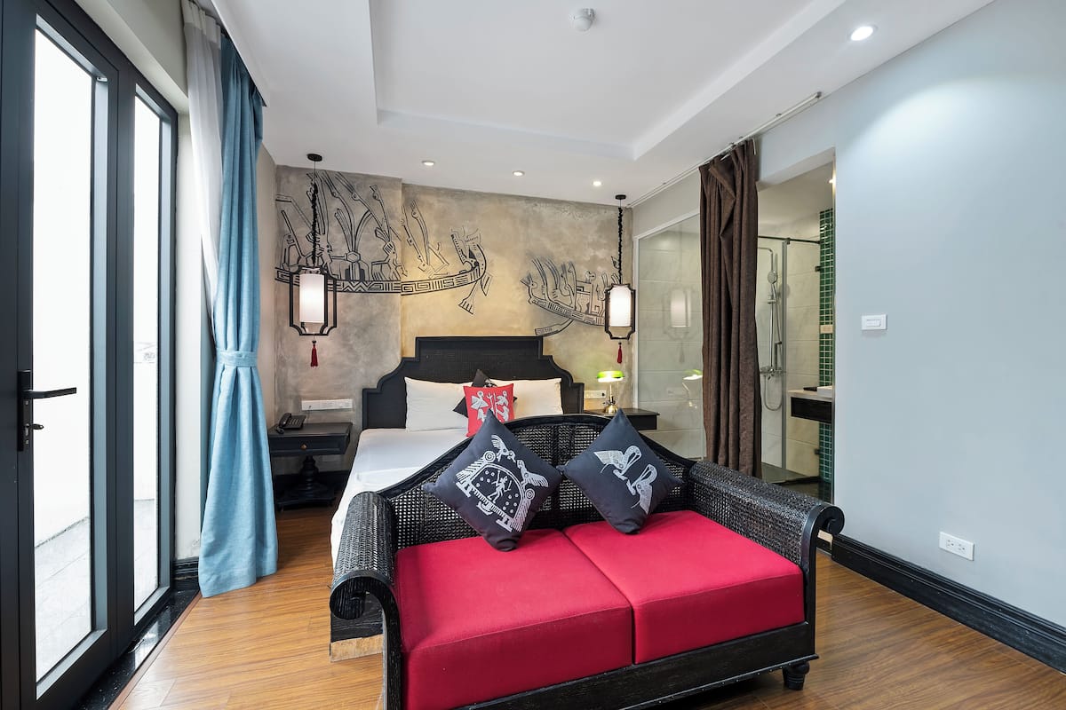 Quarto de hotel moderno com uma cama grande, um sofá vermelho e vista da cidade através de uma janela ampla.