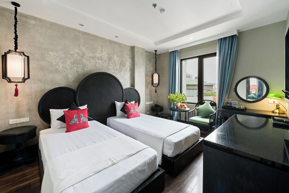 A hotel room with two beds, a desk, a chair, a small table with a plant, and a large mirror.