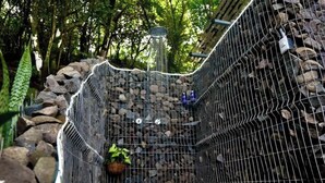 Ein Duschenbereich im Freien mit Steinwänden, umzäunt durch ein Drahtgeflecht, umgeben von Steinen und Grünpflanzen.