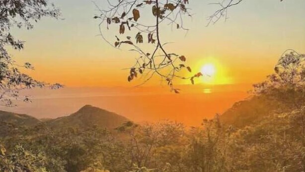 Eine Person sitzt auf einem Bett und blickt auf einen Sonnenuntergang über einer Berglandschaft.
