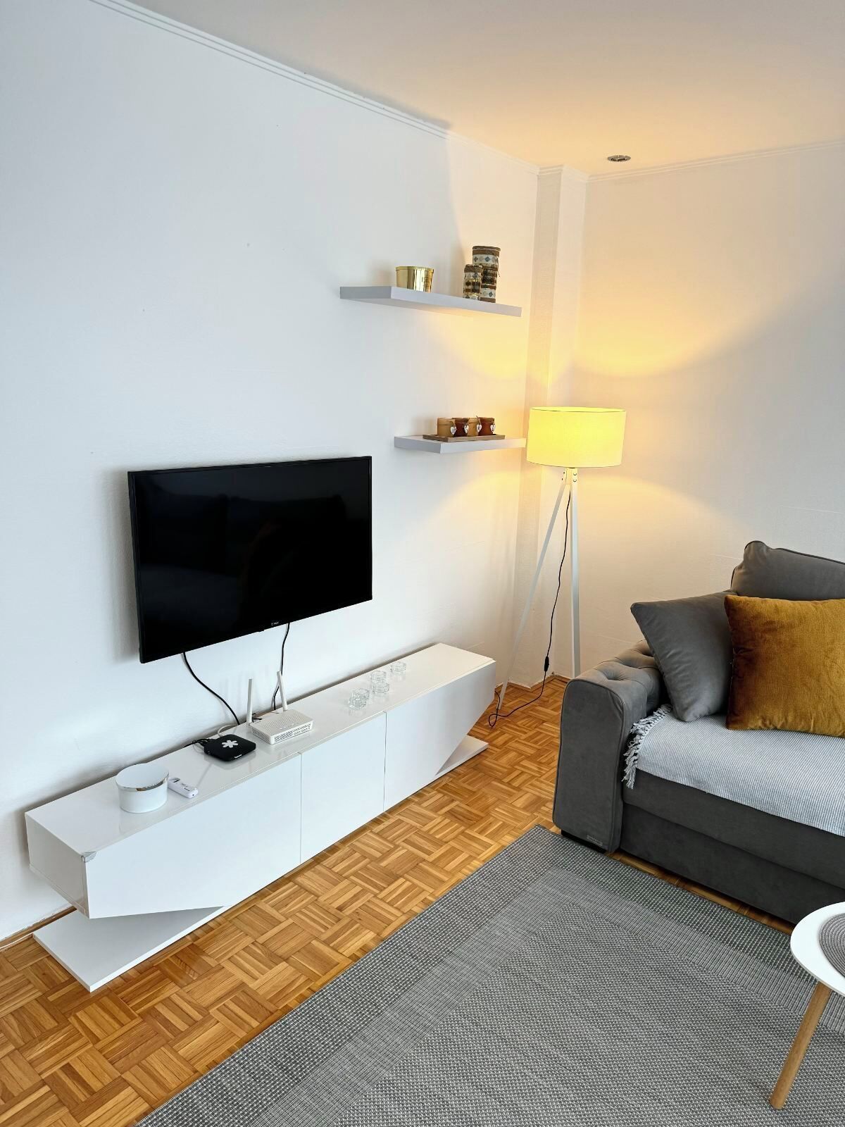 Ett modernt vardagsrum med en platt-TV som är monterad på väggen, ett vitt avlastningsbord med en router och en kaffebryggare, en grå soffa med kuddar, en golvlampa och en mönstrad matta.