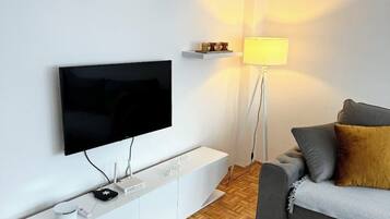 Ett modernt vardagsrum med en platt-TV som är monterad på väggen, ett vitt avlastningsbord med en router och en kaffebryggare, en grå soffa med kuddar, en golvlampa och en mönstrad matta.