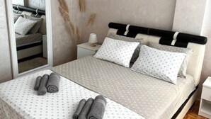 Una cama bien tendida con una colcha estampada, dos toallas enrolladas, una mesita de noche con una lámpara y un espejo que refleja la cama.