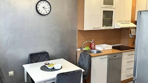 Cocina compacta con armarios blancos, un lavamanos y un lavavajillas. Hay un reloj en la pared y una mesa de comedor con sillas.