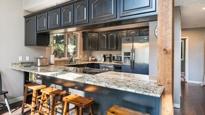 Une cuisine moderne avec des meubles de couleur sombre, un grand îlot central doté de tabourets, des appareils électroménagers en acier inoxydable et une vue sur l’extérieur.