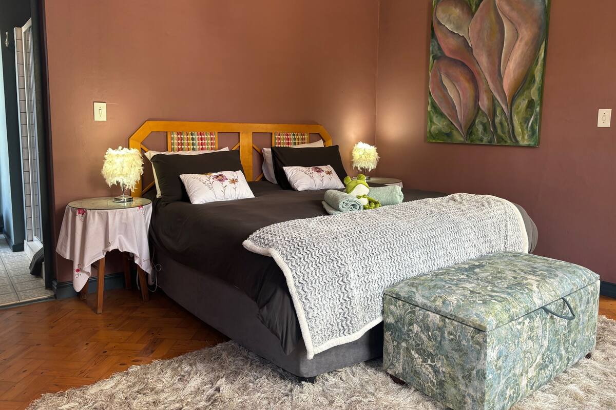 A bedroom with a large bed, a side table, a chair, a footstool, and a painting on the wall.