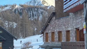 A rustic building with stone walls and wooden balconies, a snow-covered landscape, and a person skiing in the distance. - Apparemment sur les Pistes de ski Avec Piscine Risoul1850 Hautes Alpes (Risoul)
