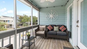 A covered porch with a ceiling fan, a wall-mounted compass, a dark-colored sofa with striped pillows, and a small table with a potted plant. - Updated Sunset Beach home w/ a 2-minute walk to the beach.  Parking for 5 cars. (Sunset Beach)