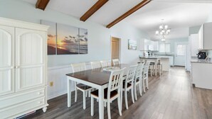 A spacious dining area with a wooden table and white chairs, a kitchen with white cabinets, and a view of the sunset through a window. - Updated Sunset Beach home w/ a 2-minute walk to the beach.  Parking for 5 cars. (Sunset Beach)