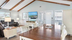 A spacious living room with a wooden ceiling, a large flat-screen TV mounted on the wall, a dining table with chairs, a sofa, and a small cabinet. - Updated Sunset Beach home w/ a 2-minute walk to the beach.  Parking for 5 cars. (Sunset Beach)