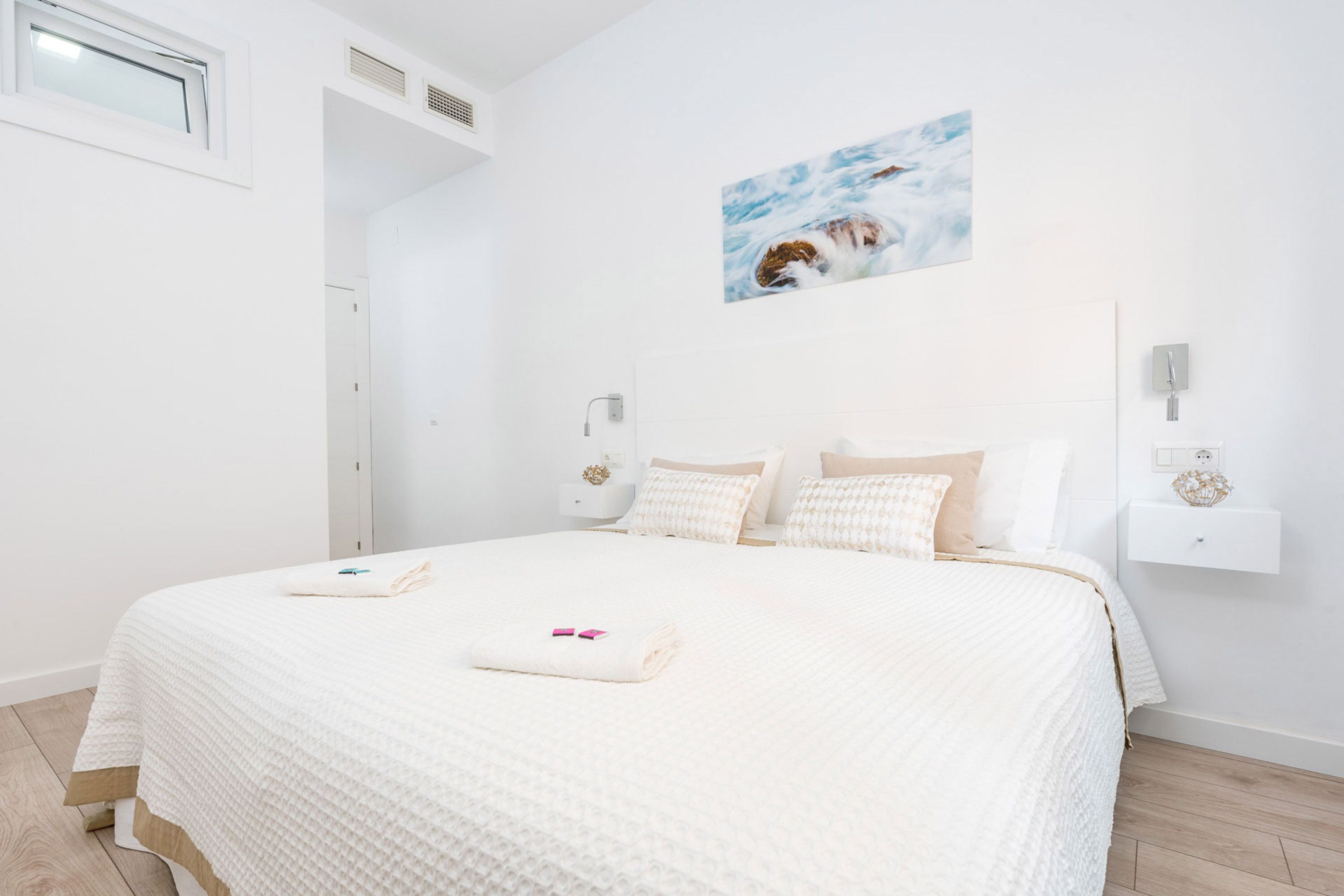 A bedroom with a large bed, white walls, a painting of a dog, and a small bedside table with a decorative item.