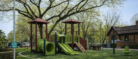 A playground with slides and climbing structures, surrounded by trees and a brick building.