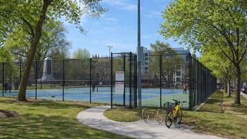 A fenced tennis court with a bicycle parked nearby, surrounded by trees and a sidewalk.