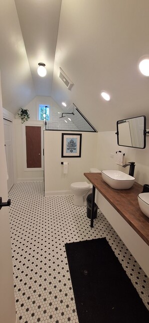 A modern bathroom with a white and black tiled floor, a wooden vanity, a white sink, and a mirror. - The Oak Park Loft (Oak Park)