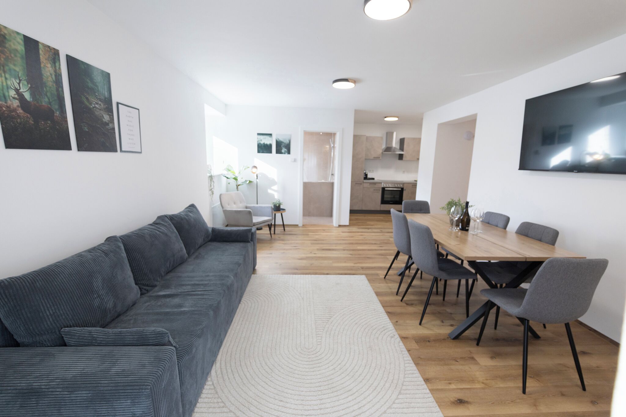 Ein modernes Wohnzimmer mit einer grauen Couch, einem Esstisch mit Stühlen und einer Küche im Hintergrund.