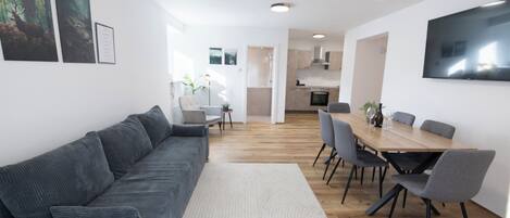 Ein modernes Wohnzimmer mit einer grauen Couch, einem Esstisch mit Stühlen und einer Küche im Hintergrund.