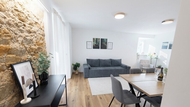 A modern living room with a stone wall, a grey sofa, a wooden dining table with chairs, and a white rug. - Modern Apartment Milo 3 room apartment for up to 6 people (Küssaberg)